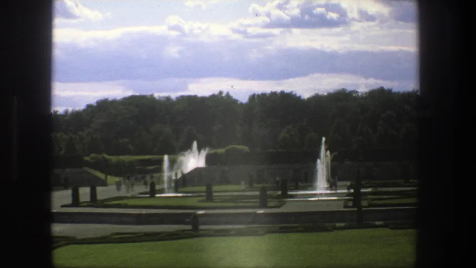 1975 8mm Stockholm park view through a dark frame, showing fountains, clipped hedges, lawns, paths, and distant visitors.
