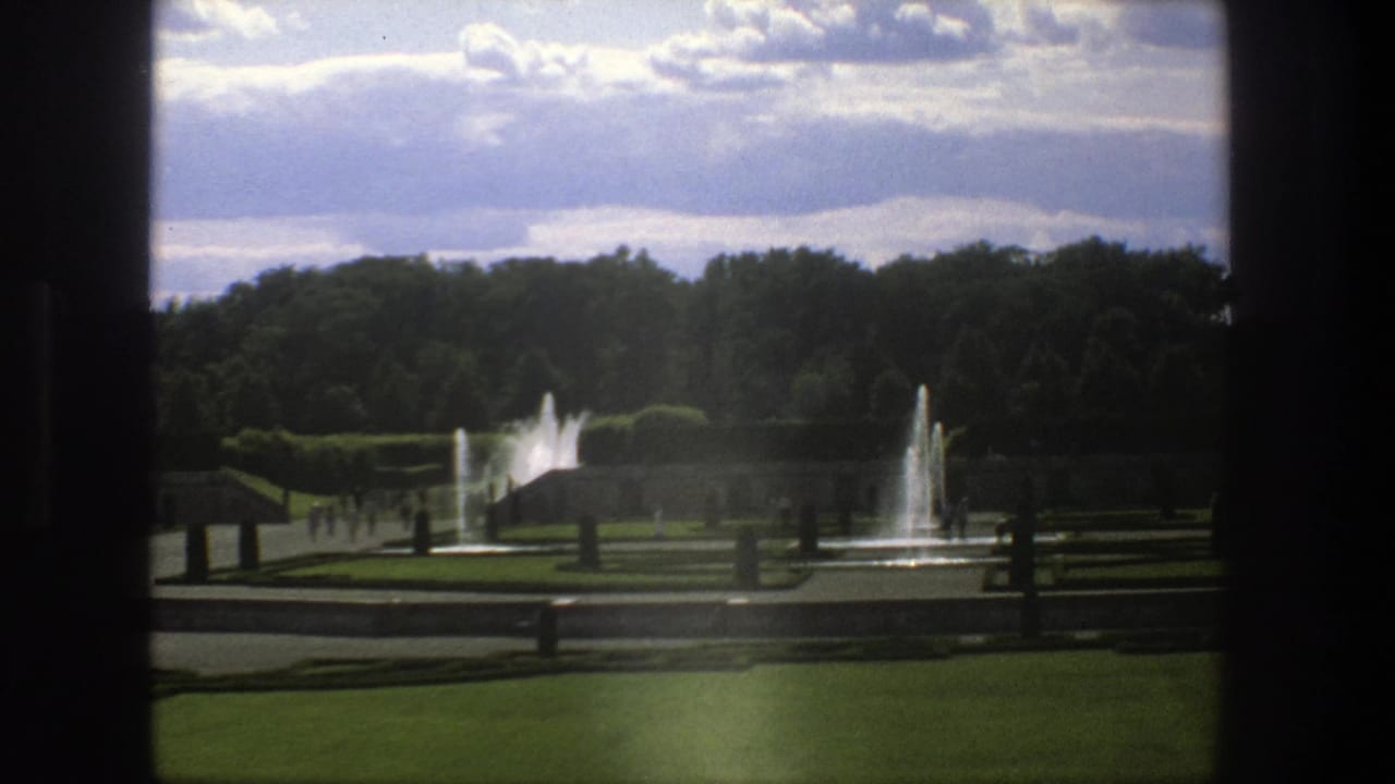 Stockholm 1975 home-movie frame of a formal park garden with multiple fountains, dark framing, paths, and tiny strolling visitors.
