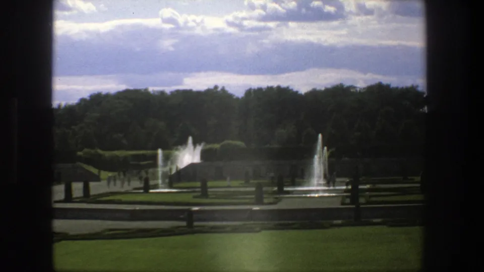 Stockholm 1975 home-movie frame of a formal park garden with multiple fountains, dark framing, paths, and tiny strolling visitors.