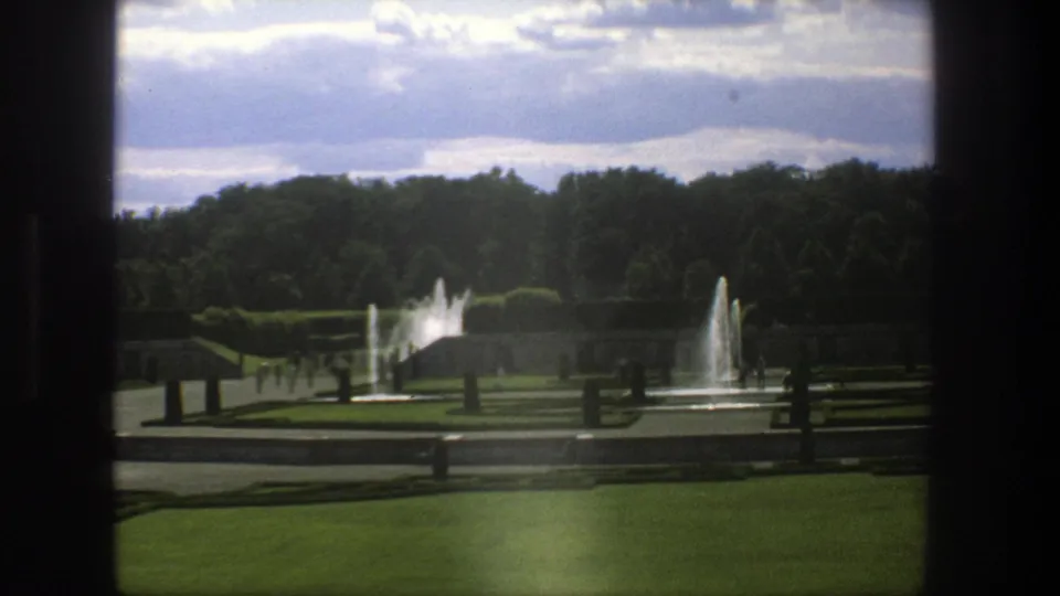 Backlit 1975 Stockholm park view with fountains, clipped hedges, lawns, tree line, and dark vertical borders from the filming position.