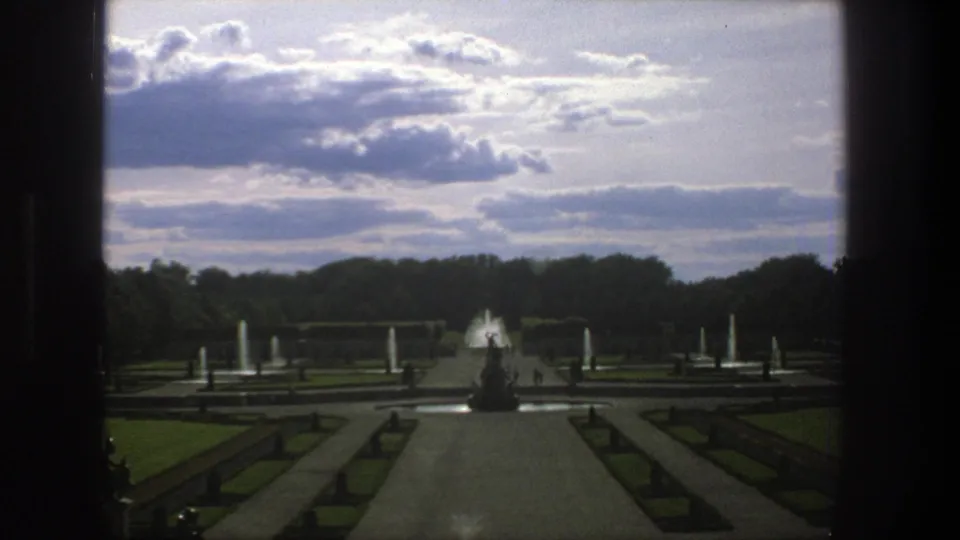 Wide 1975 Stockholm park view with a central walkway, fountain jets, formal hedges, lawns, distant visitors, and layered clouds.