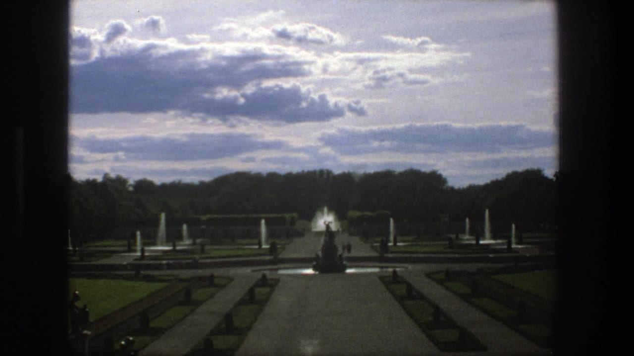 1975 Stockholm garden view with a central path, fountain jets, a statue silhouette, lawns, trees, and a cloudy sky.