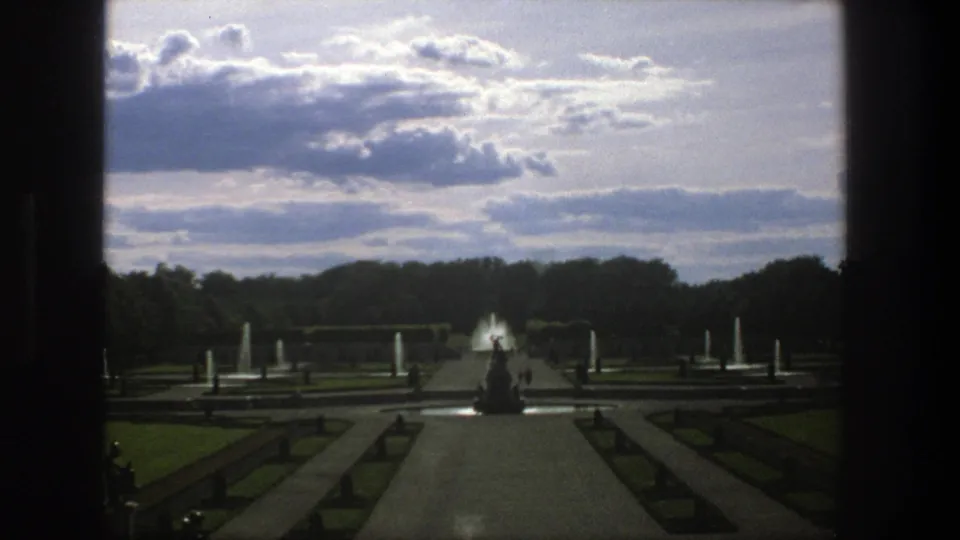 1975 Stockholm garden view with a central path, fountain jets, a statue silhouette, lawns, trees, and a cloudy sky.