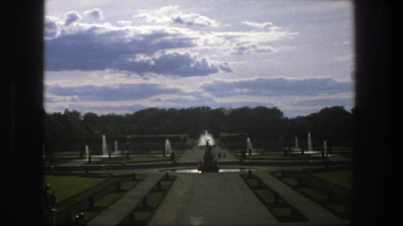 Formal garden seen through a dark frame with a central path, fountains, hedges, trees, and cloudy sky.