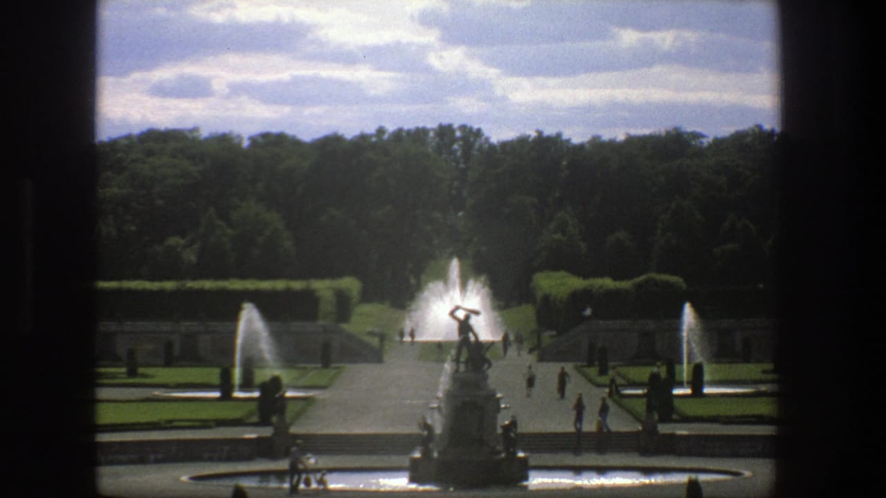 1975 Stockholm garden view with a statue fountain in the foreground, water jets on both sides, clipped hedges, paths, and distant visitors.