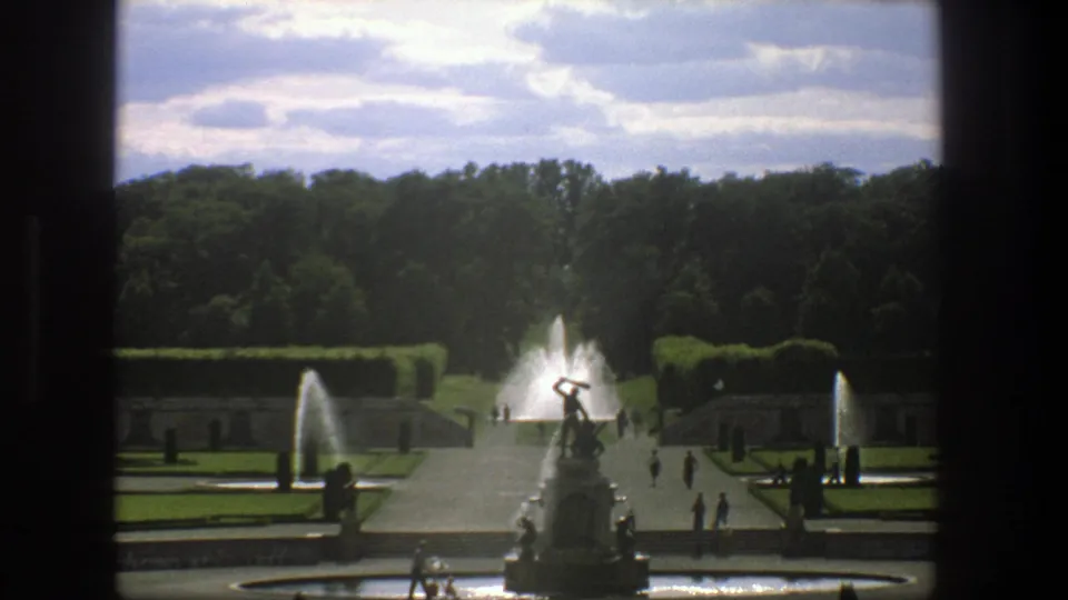 1975 Stockholm 8mm frame showing a central sculptural fountain, flanking water jets, clipped hedges, and small visitor figures in the garden.