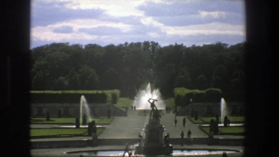 1975 Stockholm home-movie frame centered on a sculptural fountain, with symmetrical garden paths, hedges, water jets, and distant visitors.