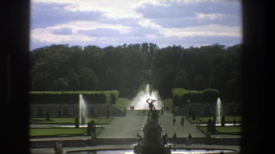 1975 Stockholm garden footage with a central fountain view, trimmed hedges, lawns, tree line, and visitors crossing the paved paths.