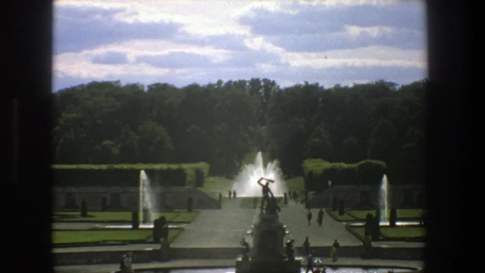 Stockholm 8mm home-movie frame from 1975 showing a formal garden axis with fountain spray, statue base, hedges, and strolling visitors.