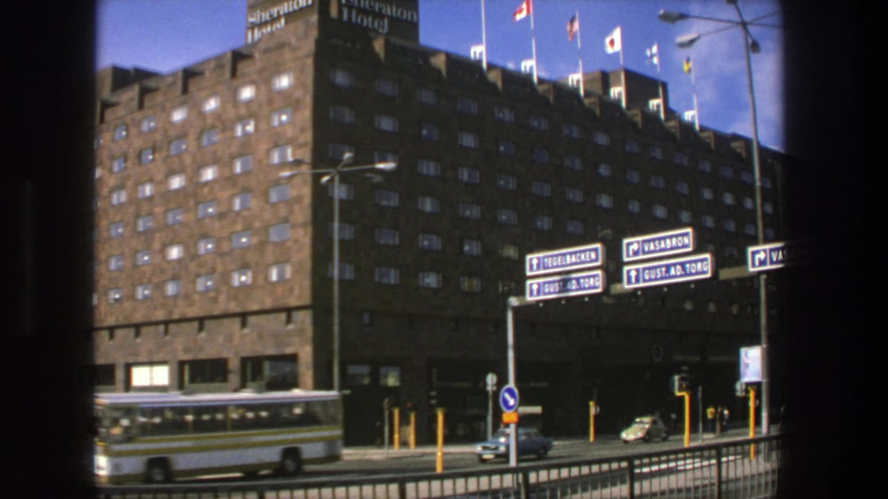 Large hotel facade with flags, street signs, a passing bus, and cars.