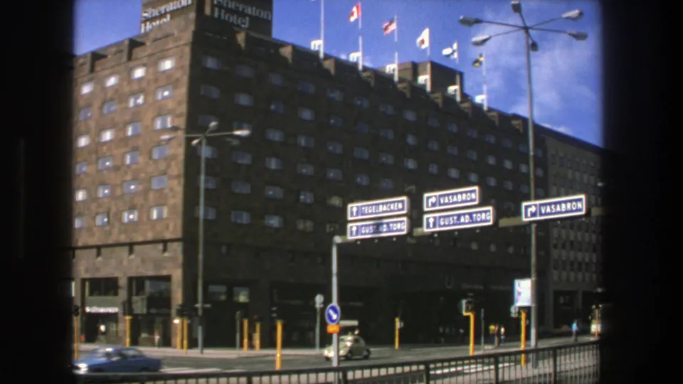 1975 Stockholm street scene with the Sheraton Hotel, rooftop flags, blue direction signs, cars, traffic lights, and railings.