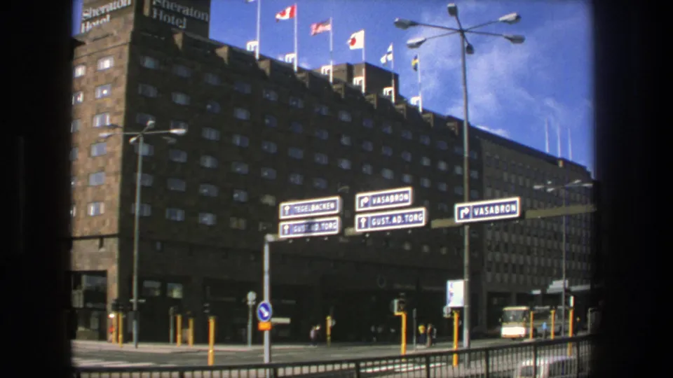 1975 Stockholm street view of the Sheraton Hotel exterior with rooftop flags, directional signs, railings, traffic lights, and a yellow-white bus.