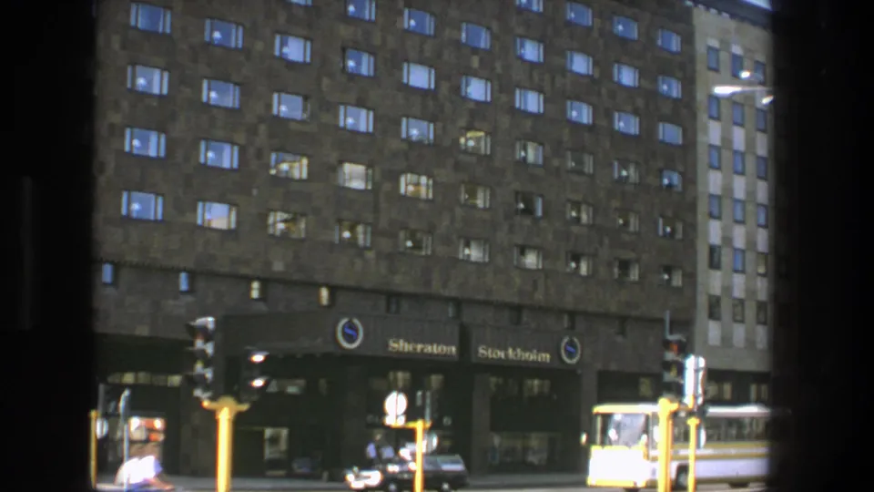 1975 Stockholm view of the Sheraton Hotel lower facade, showing lit windows, entrance signage, traffic lights, cars, pedestrians, and a bus.
