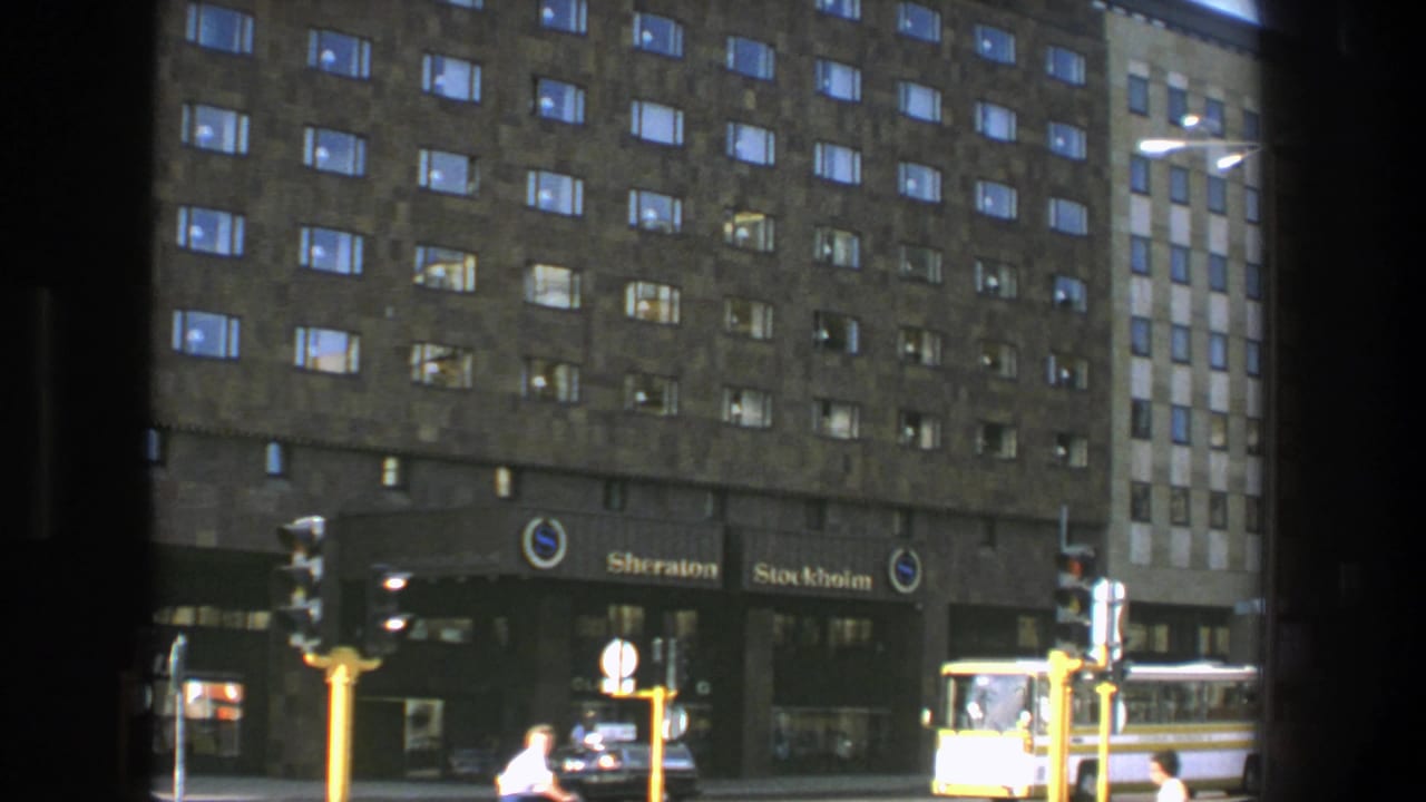 1975 Stockholm street view facing the Sheraton Stockholm, with lit windows, pedestrians, traffic signals, a car, and a yellow bus.