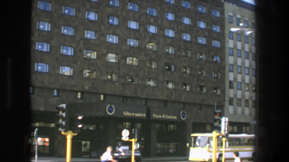 1975 Stockholm street view facing the Sheraton Stockholm, with lit windows, pedestrians, traffic signals, a car, and a yellow bus.