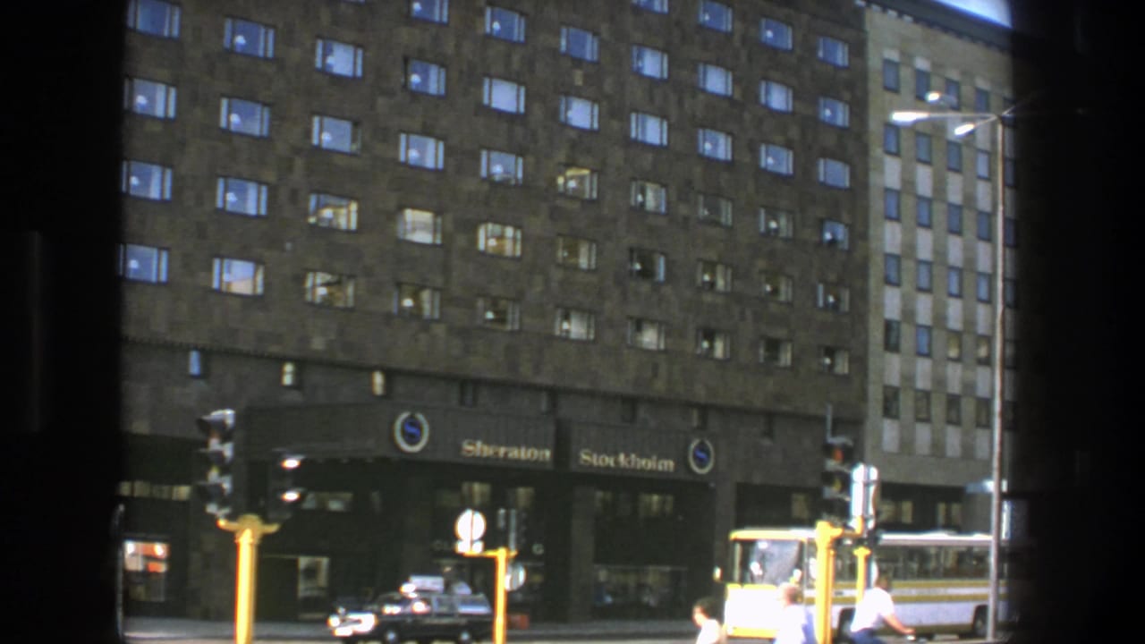 Hotel frontage with Sheraton Stockholm signs, traffic signals, pedestrians, a car, and bus traffic.