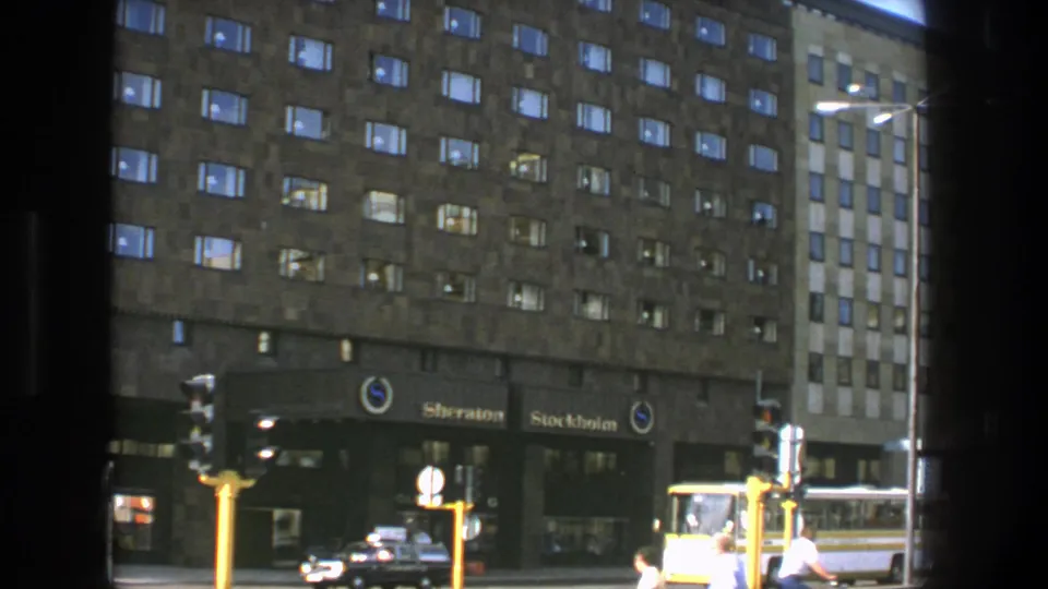 1975 Stockholm street scene outside the Sheraton Stockholm, showing hotel signage, pedestrians, a dark car, and a yellow bus near traffic lights.