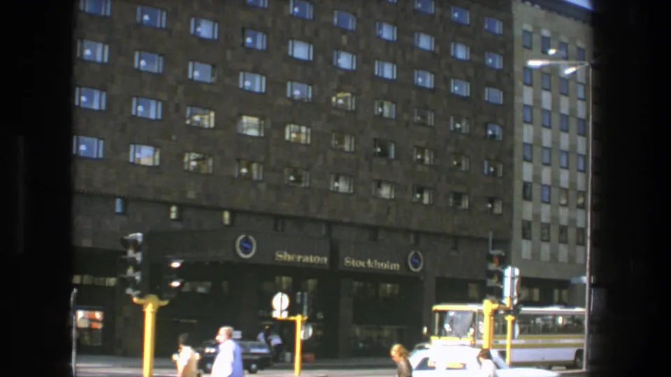 1975 Stockholm hotel-frontage scene showing the Sheraton Stockholm signs, pedestrians crossing the frame, a car, bus, and traffic lights.