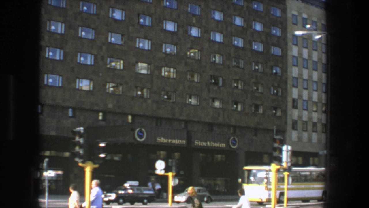 1975 Stockholm street view of the Sheraton Stockholm entrance area with pedestrians, classic cars, traffic lights, and a bus visible through a dark frame.