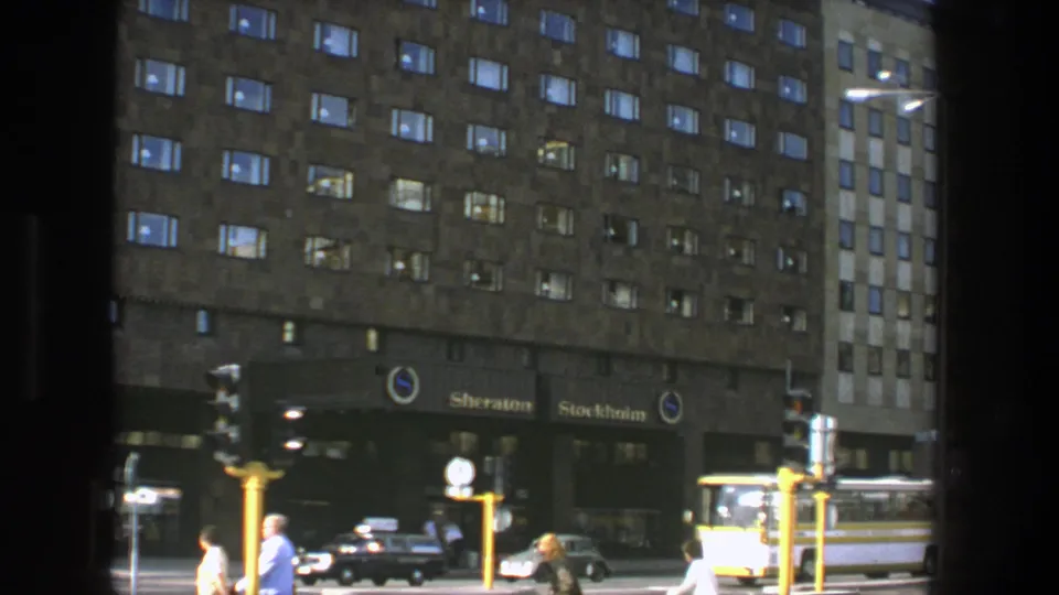 1975 Stockholm street view of the Sheraton Stockholm entrance area with pedestrians, classic cars, traffic lights, and a bus visible through a dark frame.