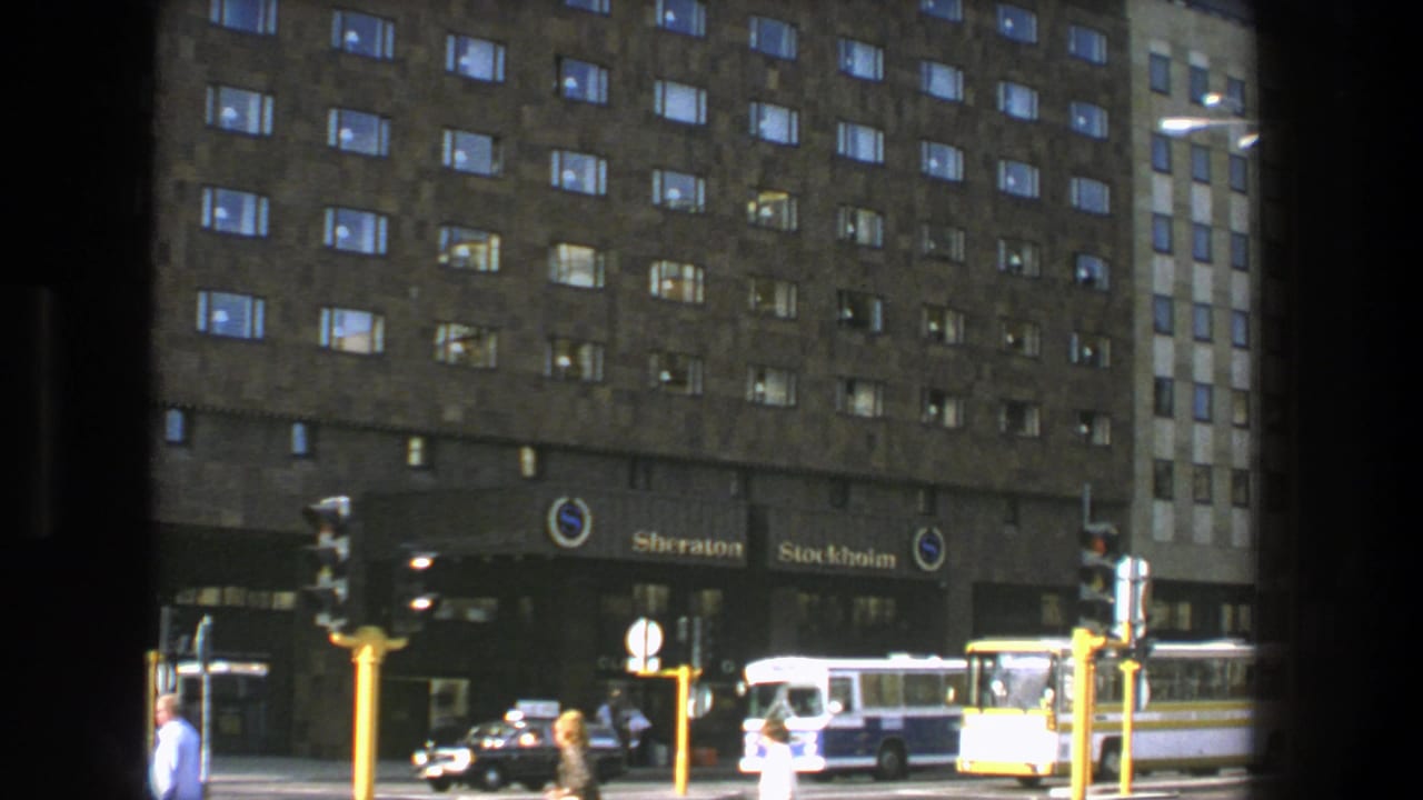 Hotel frontage with the Sheraton Stockholm sign, pedestrians, cars, a bus, and traffic lights.