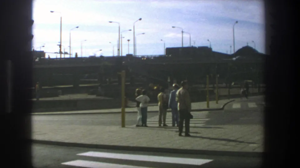 1975 Stockholm street scene with pedestrians standing near a crosswalk, broad pavement, streetlights, and passing traffic infrastructure.