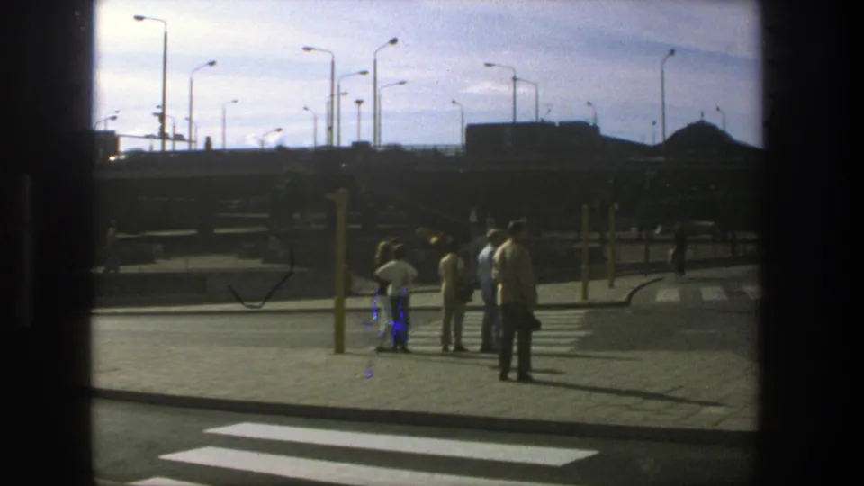 1975 Stockholm crosswalk view with pedestrians waiting on the sidewalk, streetlights overhead, and a wide roadway in the foreground.