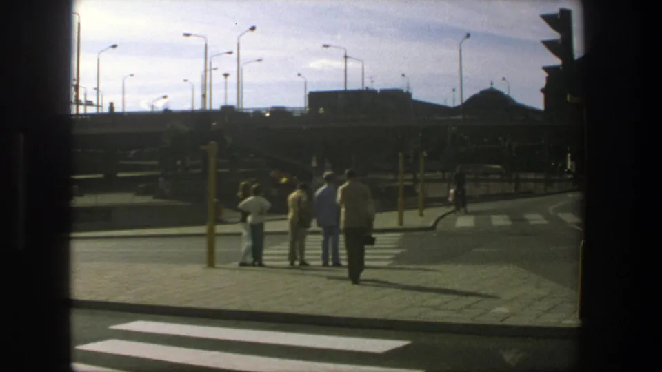 Stockholm 1975 home-movie frame of pedestrians waiting near a crosswalk beneath a bright sky and tall streetlights.