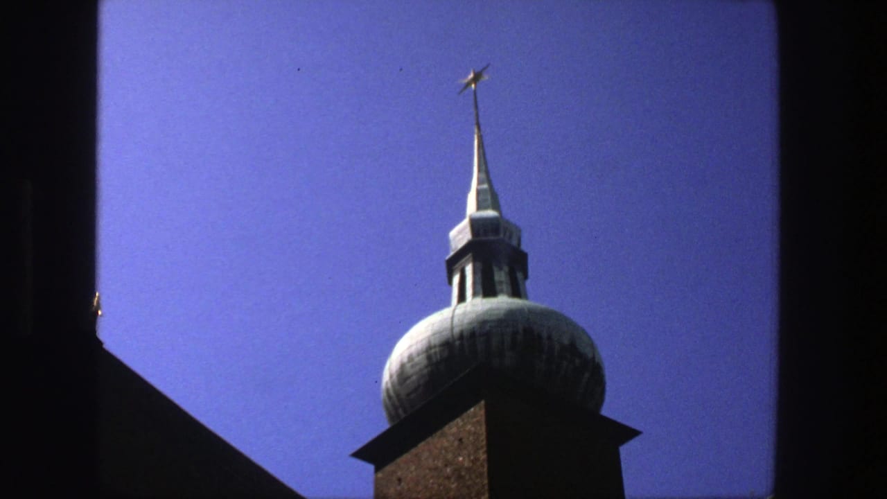 1975 Stockholm 8mm frame of a church tower cupola and star-topped spire, partly bordered by dark window trim.