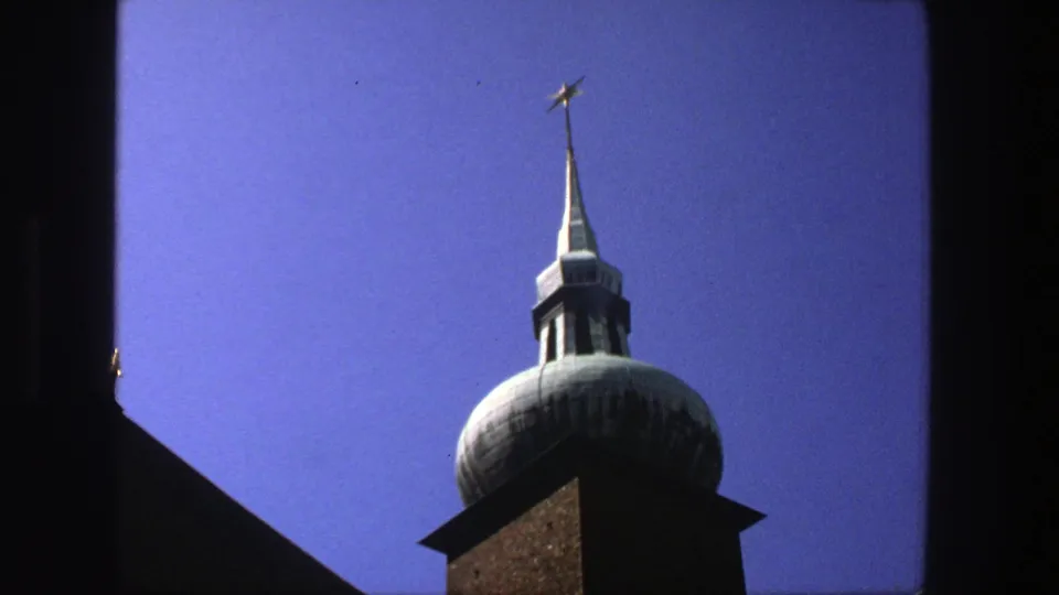 1975 Stockholm 8mm frame of a church tower cupola and star-topped spire, partly bordered by dark window trim.