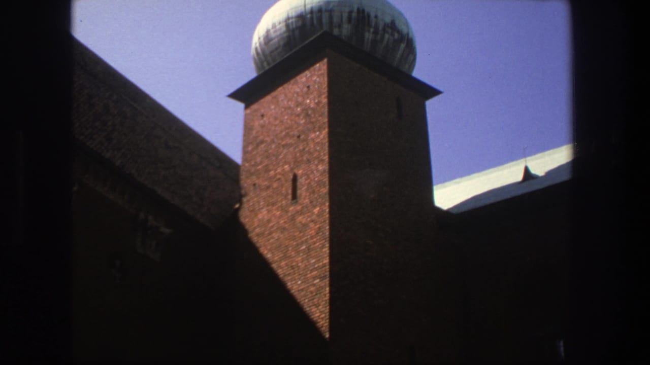 The camera holds on the Stockholm brick religious building, showing the tower, domed spire, dark roof, and blue sky through a heavy foreground frame.