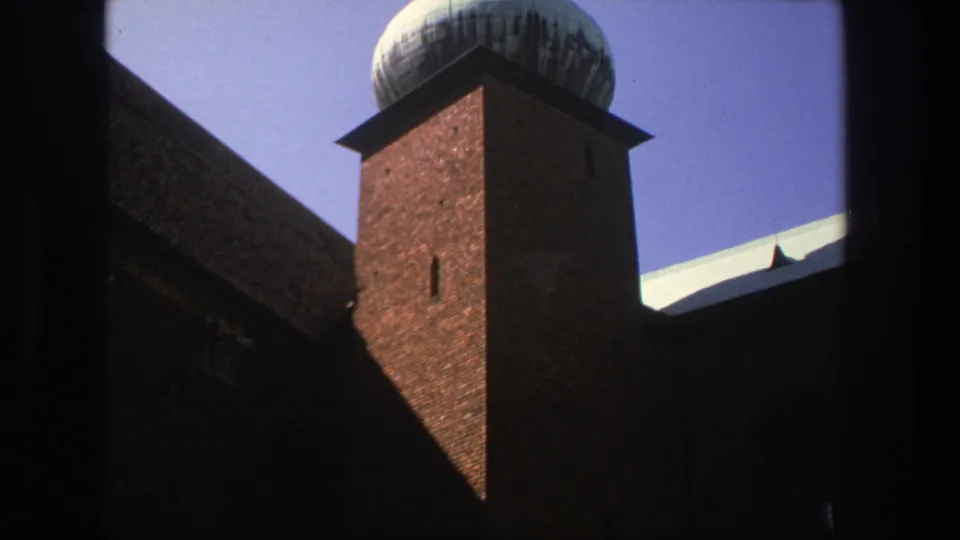 The camera holds on the Stockholm brick religious building, showing the tower, domed spire, dark roof, and blue sky through a heavy foreground frame.