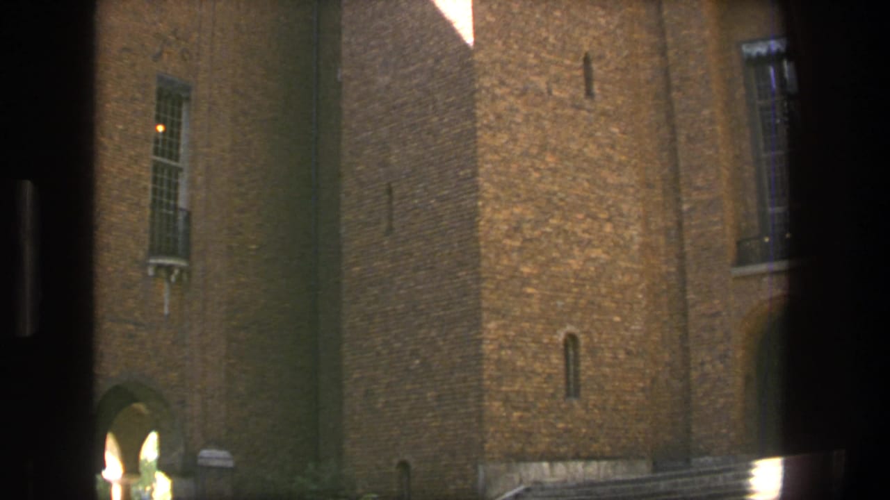 1975 Stockholm 8mm frame showing a brown brick building facade with barred windows, an arched passage, and strong sunlight across the masonry.