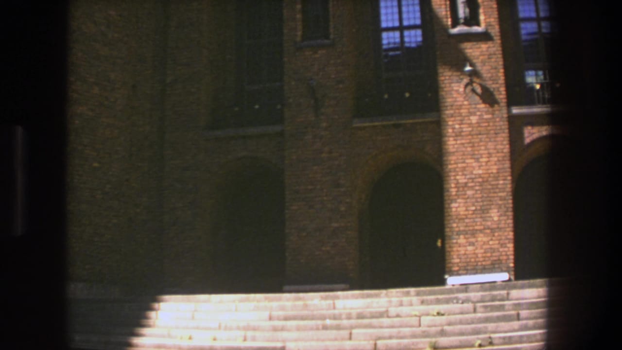 1975 Stockholm frame with broad stone steps leading toward a brown brick exterior, arched doorway, and tall windows partly crossed by shadow.