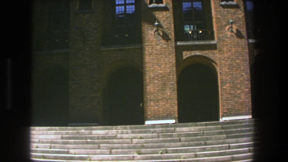 Late shot from the 1975 Stockholm clip showing repeated arched brick entrances, barred windows, and broad stone steps in warm sunlight.