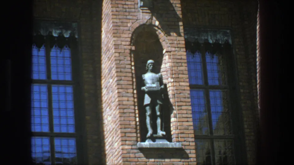 Handheld 8mm view in Stockholm of a standing figure in a brick recess next to tall multi-paned windows with blue reflections.