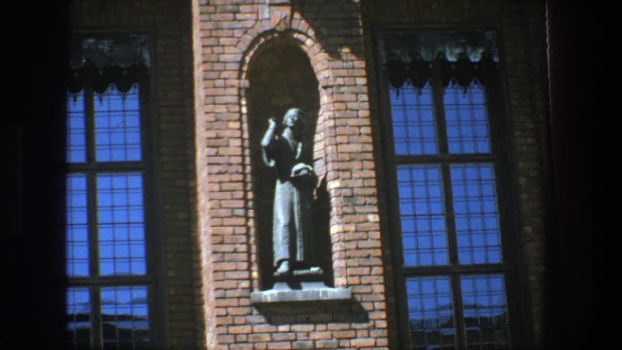 1975 Stockholm 8mm frame showing a robed statue in a brick niche between tall blue-reflecting windows.