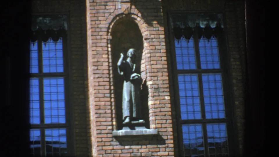 1975 Stockholm 8mm frame showing a robed statue in a brick niche between tall blue-reflecting windows.