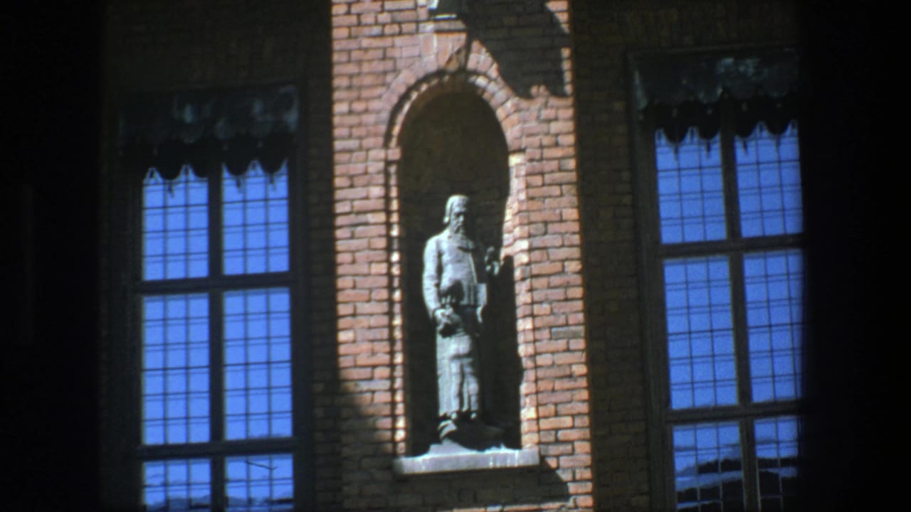 1975 Stockholm 8mm frame with a standing statue set into a brick facade near blue-paned windows and deep shadows.