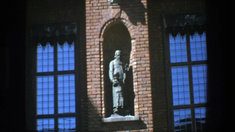1975 Stockholm 8mm frame with a standing statue set into a brick facade near blue-paned windows and deep shadows.