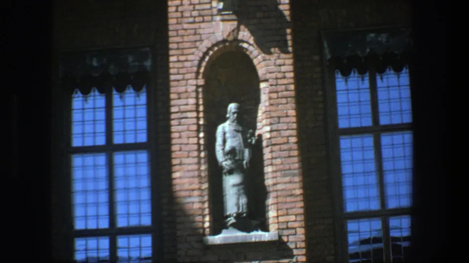 1975 Stockholm 8mm frame showing a brick facade, arched statue niche, blue window panes, and heavy shadow across the wall.