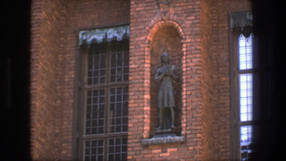 1975 Stockholm 8mm frame with a closer view of a dark robed statue recessed in brick beside tall gridded windows.