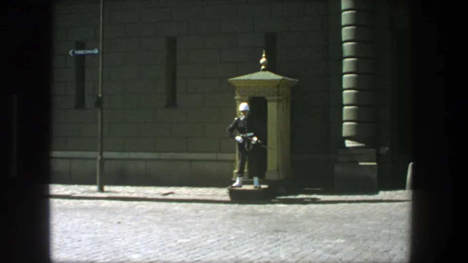 1975 Stockholm frame holding on a white-helmeted guard beside a sentry box, with cobblestones and formal stone architecture visible.
