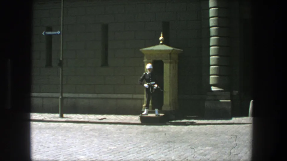 Wide 1975 Stockholm 8mm frame showing a ceremonial guard with a rifle beside an ornate sentry box and stone building.