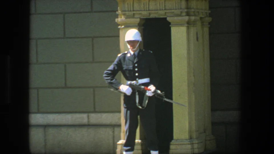 Closer 8mm view of the Stockholm ceremonial guard in dark uniform, white helmet and gloves, holding a rifle at the post.
