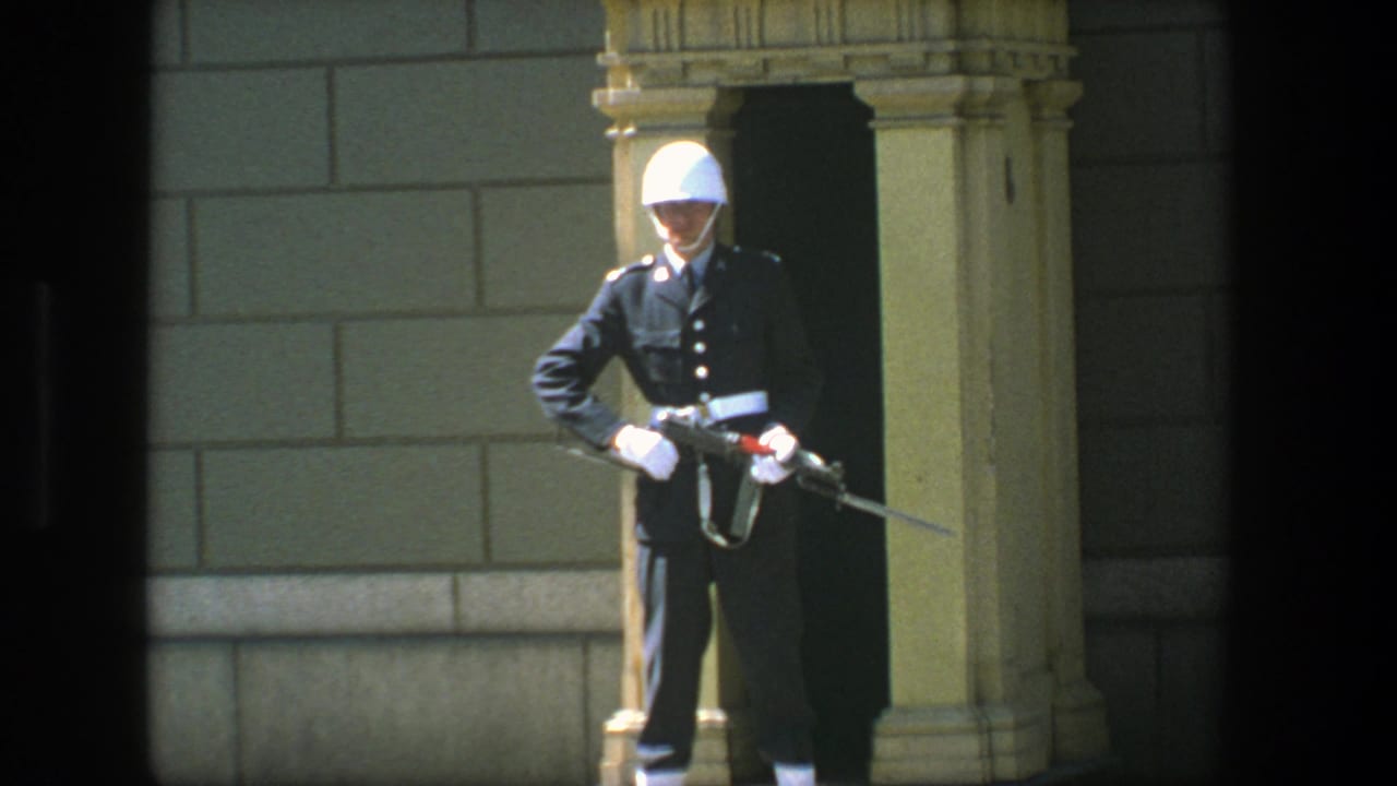 1975 Stockholm home-movie frame showing a ceremonial guard in dark dress uniform and white helmet holding a bayonet-fitted rifle.