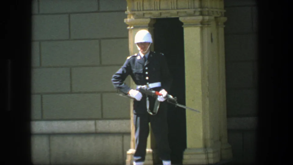 1975 Stockholm home-movie frame showing a ceremonial guard in dark dress uniform and white helmet holding a bayonet-fitted rifle.