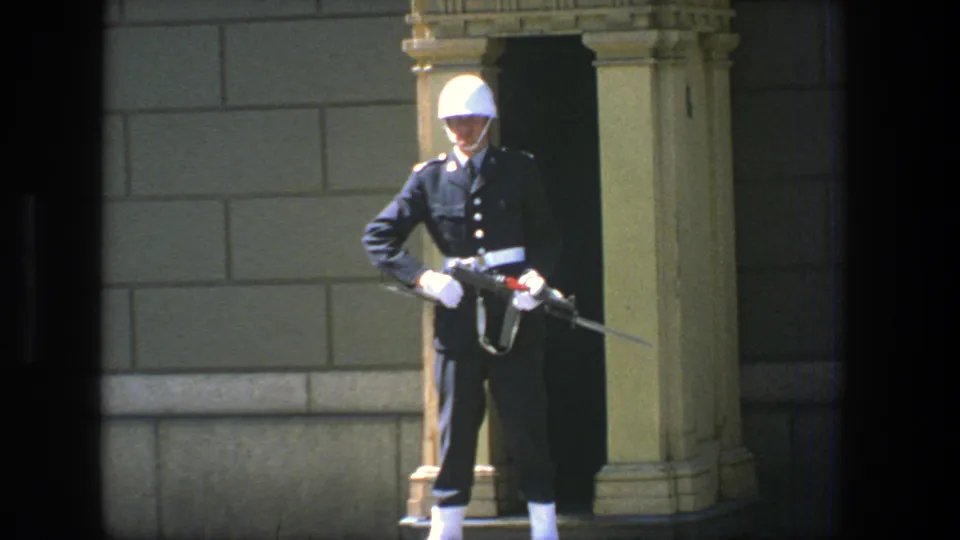 1975 Stockholm 8mm frame showing a ceremonial sentry in dark uniform, white helmet, and gloves against a stone facade.