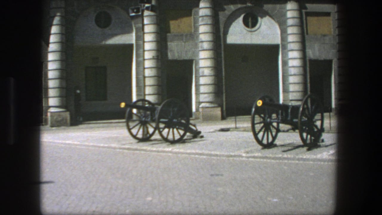 1975 Stockholm home-movie frame showing two black wheeled cannons in front of a tan stone facade with arched columns and round windows.