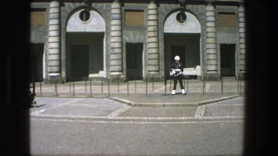 1975 Stockholm 8mm frame showing a uniformed guard with a white helmet and rifle outside a formal stone building.